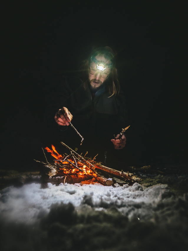 a camper tends to a fire in the snow, the image lit only by the glow of the fire and a beam of light from a headlamp